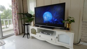 A living room with a flat-screen TV, a white entertainment unit, a potted plant, and a view through a glass door. - Rim Chaam By Roomquest (Cha-am)