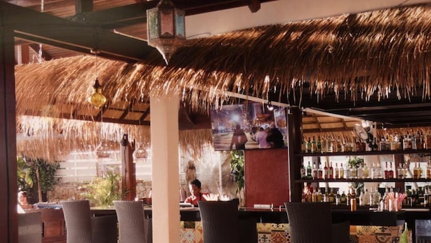 Zona de bar al aire libre con techo de paja, sillas de mimbre y una variedad de botellas en estantes.