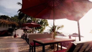 Zona de estar al aire libre frente a la playa, con mesas y sillas bajo grandes sombrillas, con vistas al océano.
