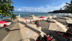 Un restaurante frente a la playa con mesas al aire libre bajo sombrillas, vistas al océano y un ambiente tropical.