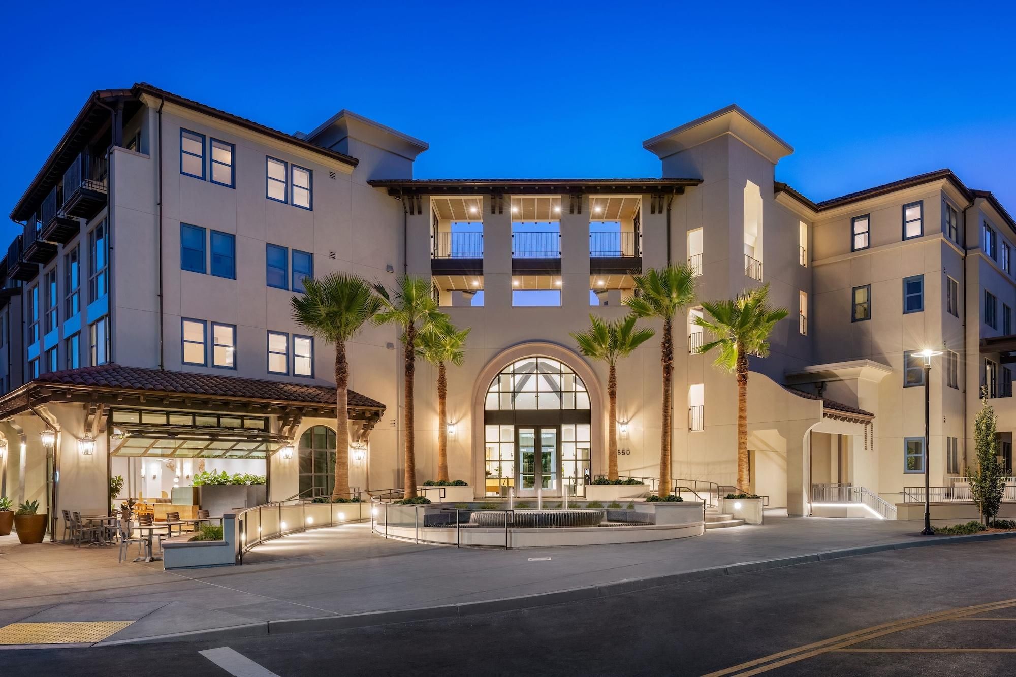A multi-story hotel with a central entrance, palm trees, and a covered outdoor seating area.