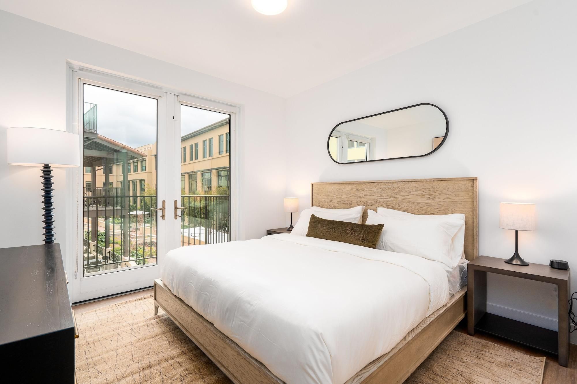 A hotel room with a bed, two bedside tables with lamps, a large mirror, and a sliding glass door leading to an outdoor area.