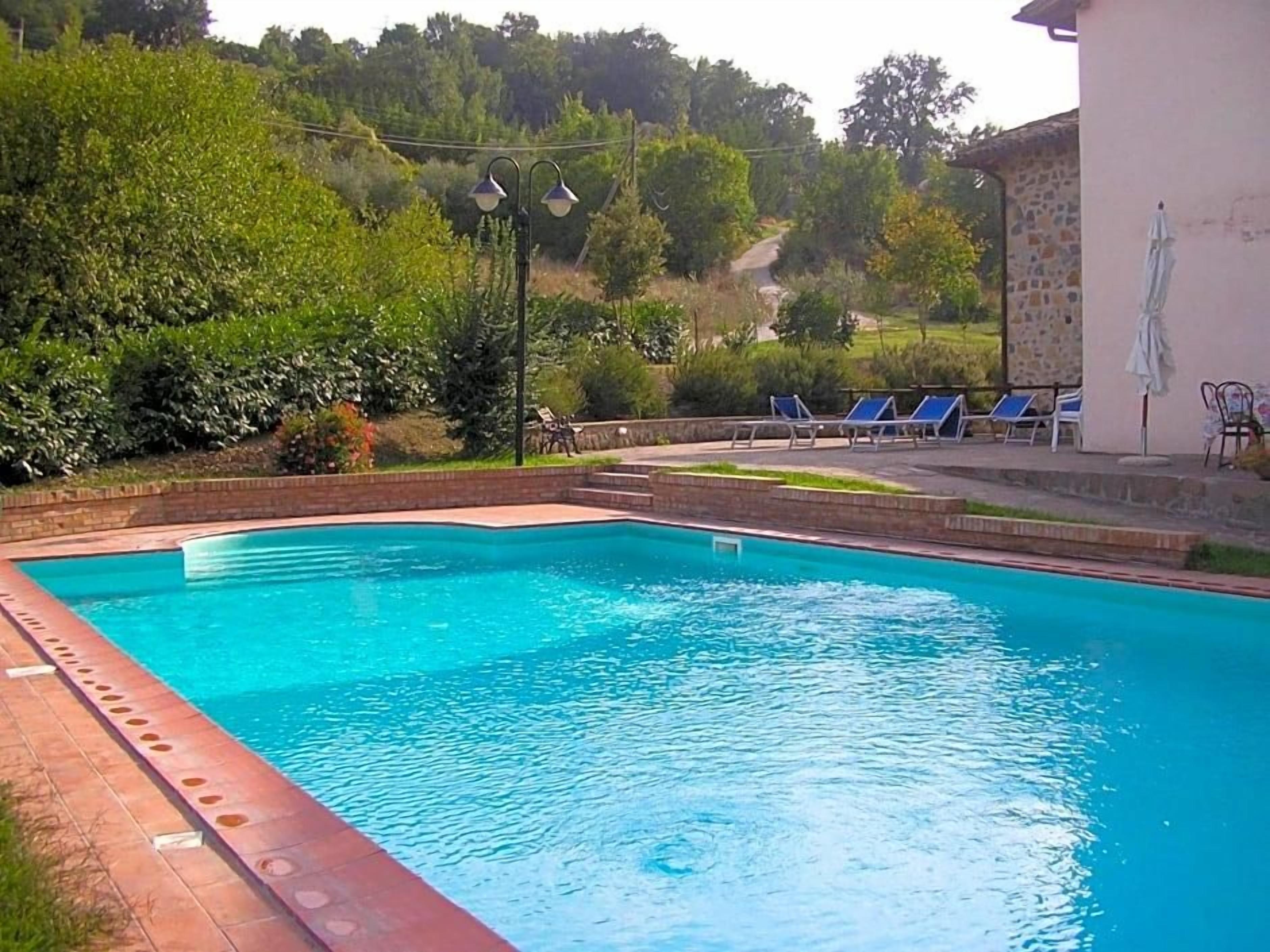 A swimming pool with a brick edge, surrounded by greenery and a building with a stone wall.