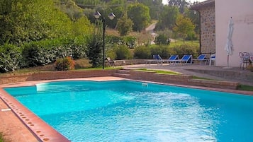 A swimming pool with a brick edge, surrounded by greenery and a building with a stone wall.