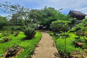 Un camí al jardí que condueix a un edifici tradicional amb teules, envoltat de vegetació exuberanta.
