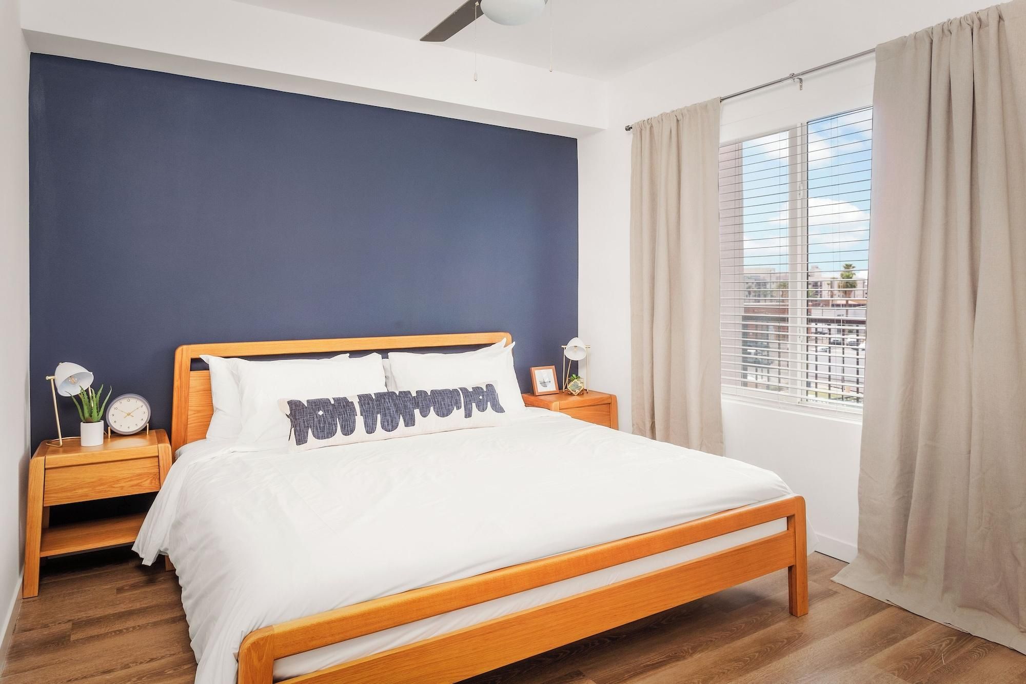 A bedroom with a wooden bed, a nightstand with a clock, a lamp, and a window with curtains.