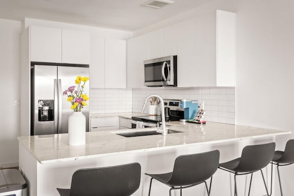 A modern kitchen with white cabinets, a stainless steel refrigerator, a microwave, a coffee machine, and a vase with flowers on the counter. - Locale Midtown Phoenix (Phoenix)