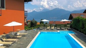 A swimming pool with lounge chairs and umbrellas, a mountain view, and a building with a balcony. - Hotel Adler (Menaggio)