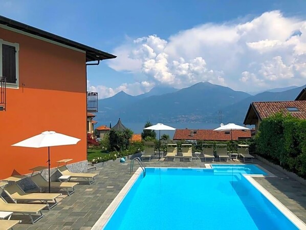 A swimming pool with lounge chairs and umbrellas, a mountain view, and a building with a balcony. - Hotel Adler (Menaggio)
