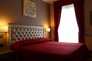 Habitación de hotel con cabecera capitonada, colcha roja, una obra de arte en la pared y ventana con cortinas rojas.