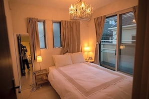 A bedroom with a bed, two bedside lamps, a chandelier, a window with curtains, and a mirror.