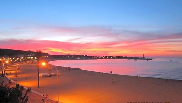 Una playa al atardecer con un mar tranquilo, una costa sinuosa y luces de la ciudad a lo lejos.