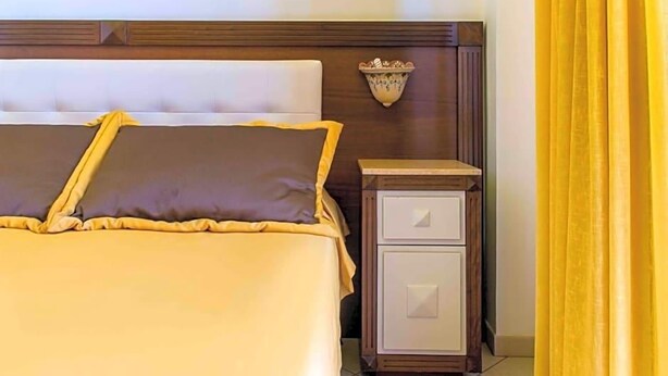 A neatly made bed with a headboard, a bedside table with drawers, and a wall-mounted light fixture. - Hotel Egitarso Sul Mare (San Vito Lo Capo)