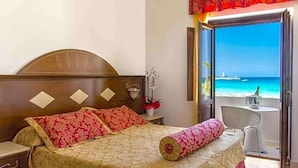 Un dormitorio con cama, mesita de noche, una pequeña mesa con lámpara y vista a la playa a través de una puerta abierta.