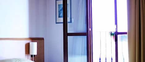 Quarto de hotel com cama, mesa de cabeceira, luminária, quadro pendurado na parede e porta de vidro corrediça que dá para uma varanda.