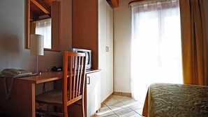 Una habitación de hotel con un escritorio de madera, una silla, una cama con colcha estampada, un televisor sobre un mueble y una ventana con cortinas.
