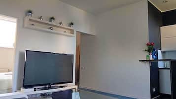 A modern living room with a flat-screen TV, a glass coffee table, a sofa, and a kitchen area with a shelf above the counter.