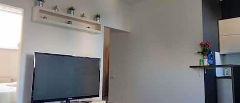 Uma sala de estar moderna com televisão de tela plana, uma mesa de centro de vidro, um sofá e uma área de cozinha com prateleira acima do balcão.