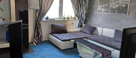 A compact room with a sofa bed, a desk, a chair, a TV, and a window with curtains.