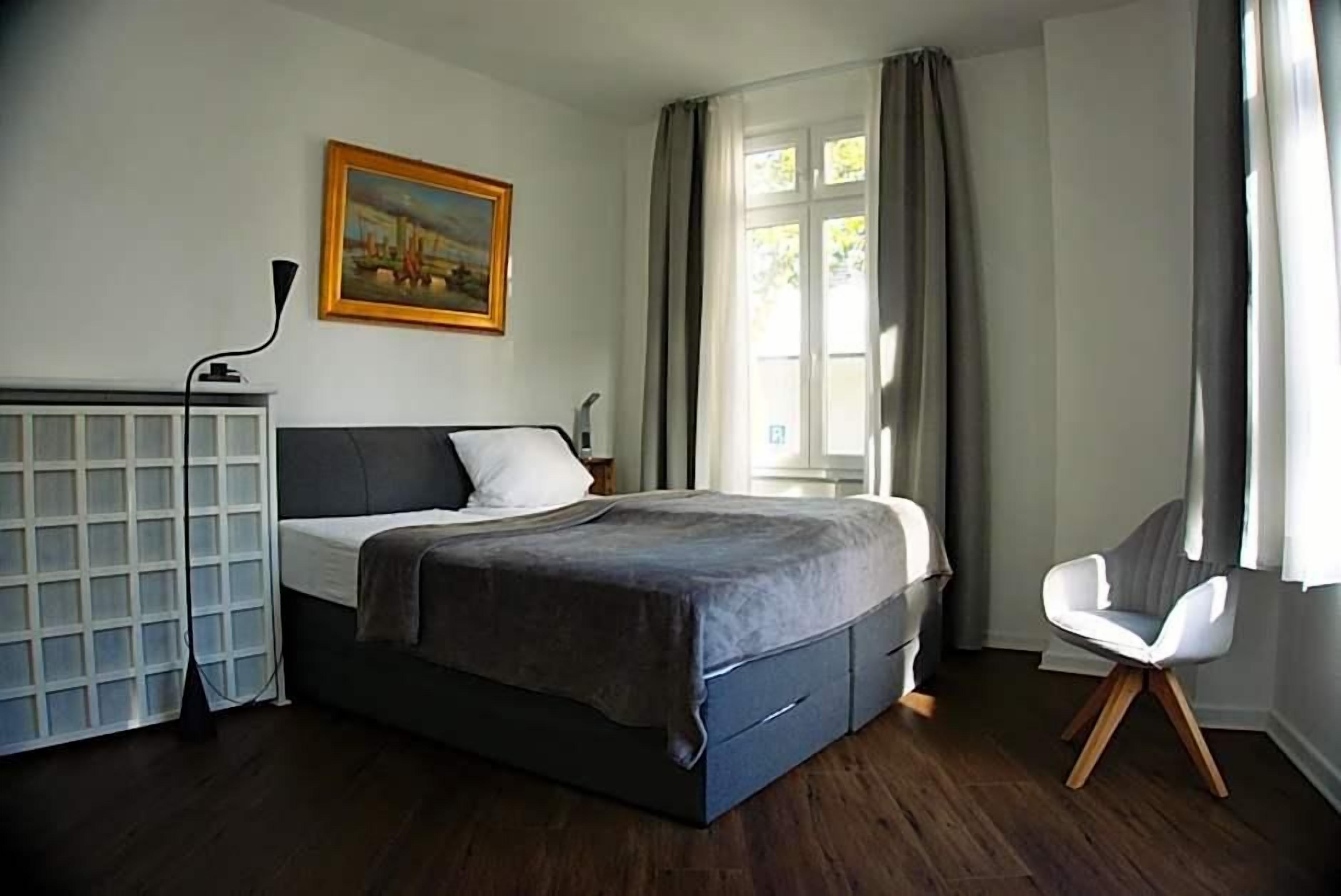 Een slaapkamer met een bed, een bureau, een stoel, een schilderij en een raam met gordijnen.