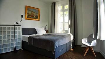 Ein Schlafzimmer mit Bett, Schreibtisch, Stuhl, einem Bild und einem Fenster mit Vorhängen.