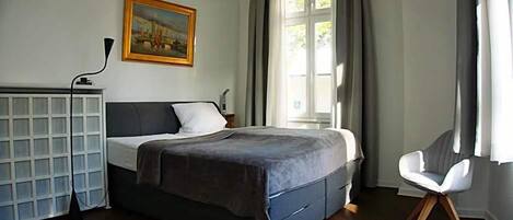 A bedroom with a bed, a desk, a chair, a painting, and a window with curtains.
