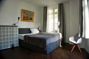 Un dormitorio con cama, escritorio, silla, un cuadro y ventana con cortinas.