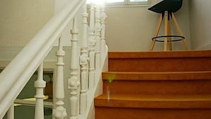 Una escalera con balaustres blancos y peldaños de madera que conduce a una habitación con ventana y una silla.
