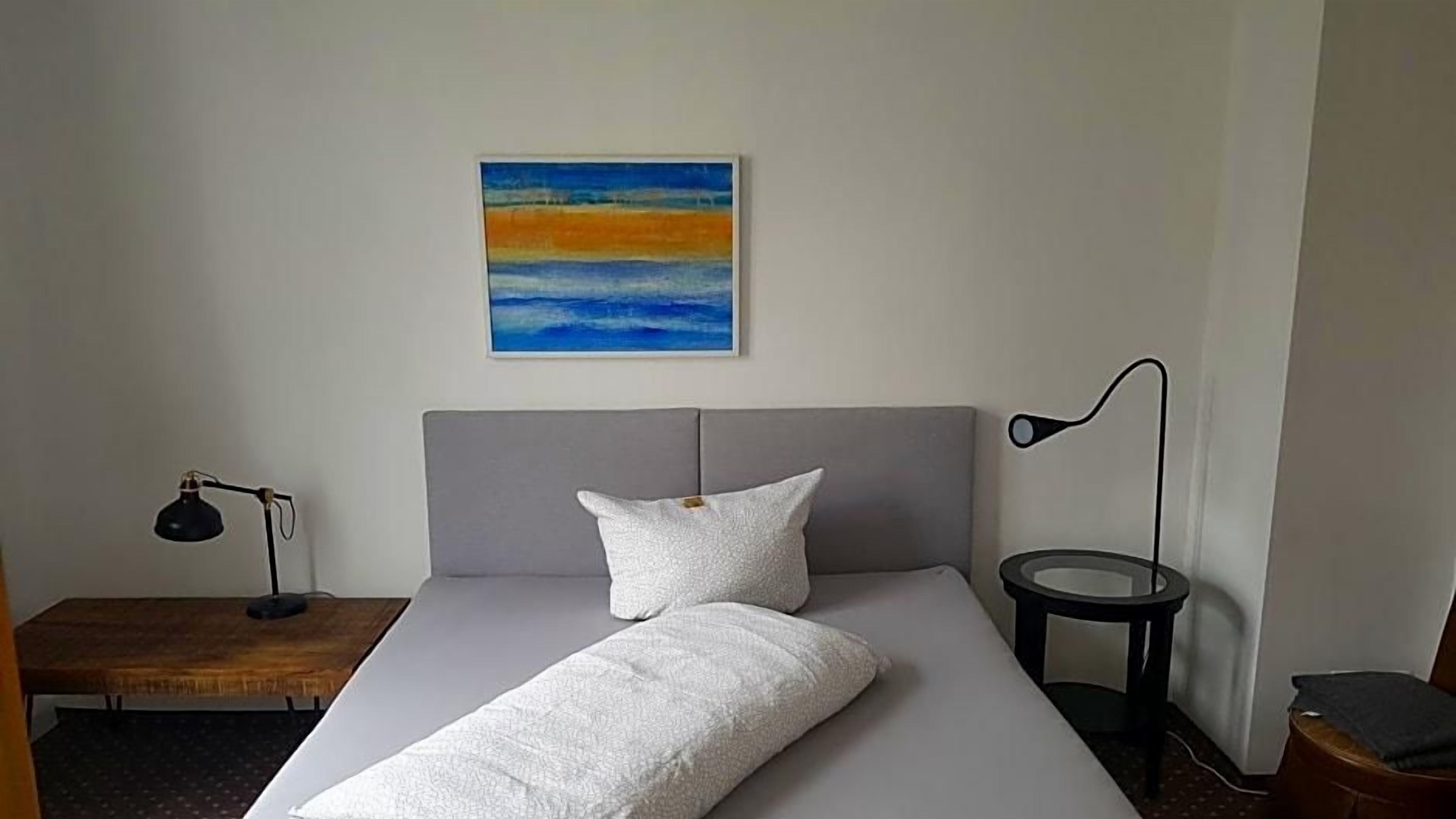 Una camera d'albergo con un letto, un comodino, una lampada, un tavolino e un quadro appeso al muro.