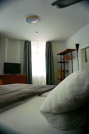 Hotellihuone, jossa on sänky, puinen lipasto, jonka päällä on televisio, ikkuna, jossa on verhot, ja hylly, jossa on vaatteita.