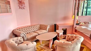 Una habitación con decoración rosa, un sofá, dos sillones, una mesita y una cama.
