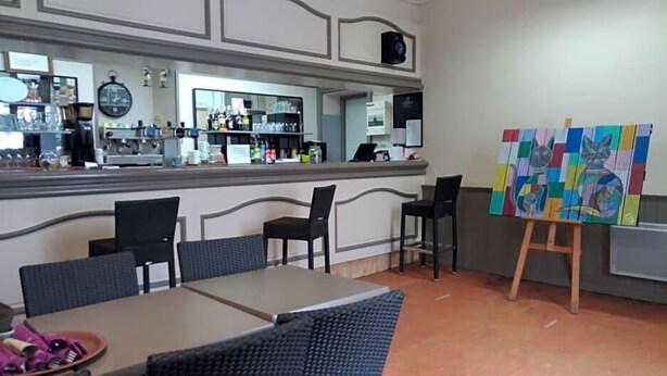A bar area with a counter, chairs, and an easel with a painting. - Le Lion D Or (Bernay)