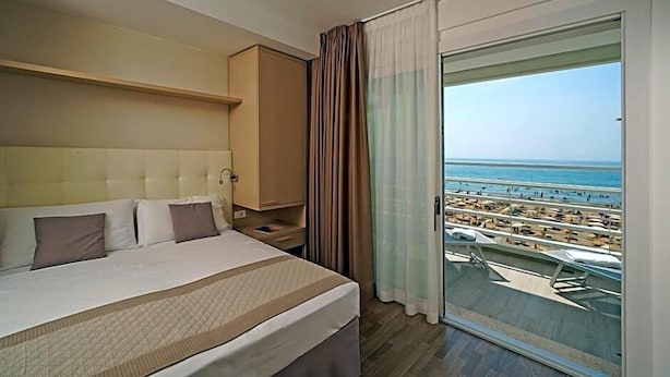 A hotel room with a large bed, a nightstand, a wardrobe, and a balcony with a view of the beach and ocean. - Hotel Strand A Frontemare (Jesolo)
