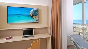 A room with a TV displaying a beach scene, a laptop on a desk, a chair, and a balcony with a view of the beach. - Hotel Strand A Frontemare (Jesolo)