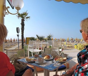 A table with breakfast items, outdoor seating, and a view of the beach. - Hotel Strand A Frontemare (Jesolo)