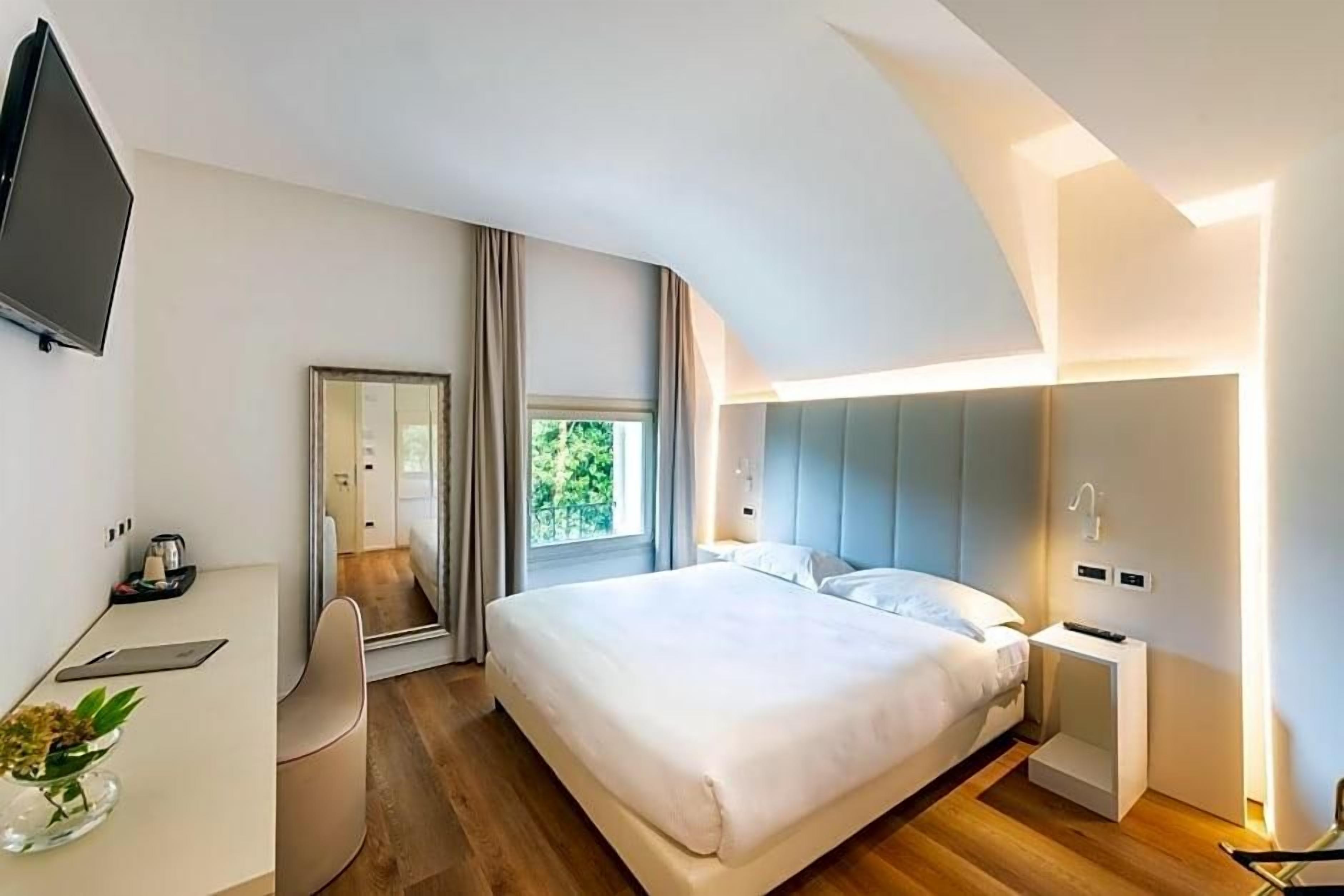 A modern hotel room with a large bed, a desk with a chair, a TV, and a window with curtains.