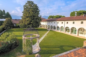 A courtyard with a gazebo, a large tree, and a building with arched windows. - Villa I Pini (Civitella in Val di Chiana)