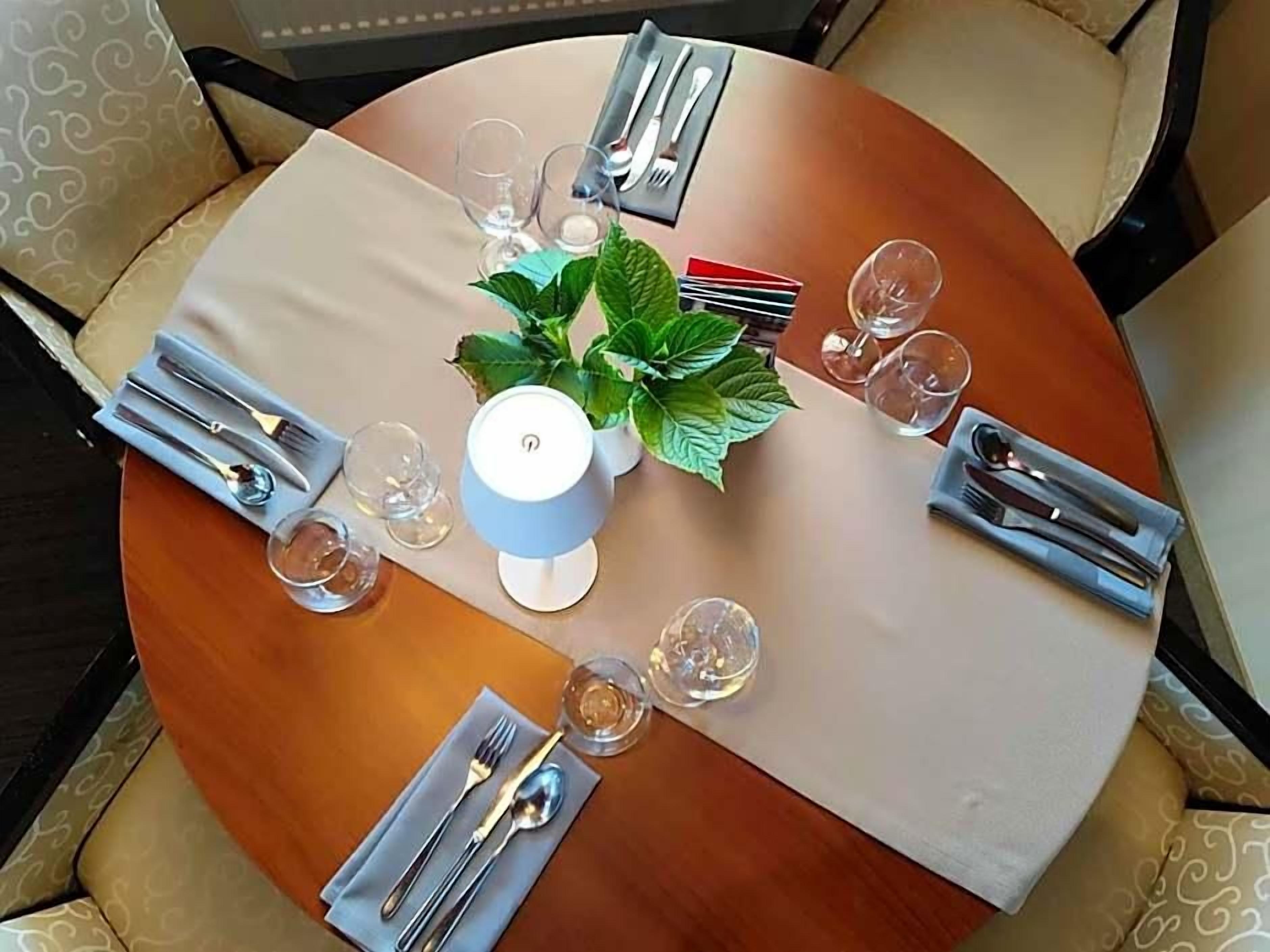 A round dining table with a white tablecloth, set with cutlery, glasses, and a potted plant.