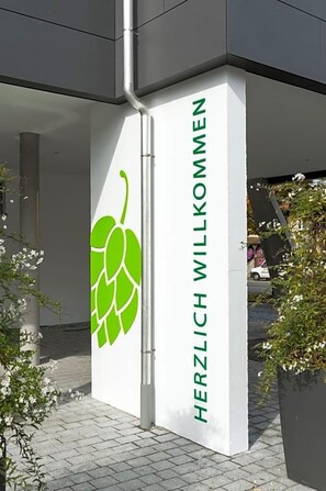 Entrada de un edificio con un cartel que dice 'HERZLICH WILLKOMMEN' y un logotipo de hoja de lúpulo verde.