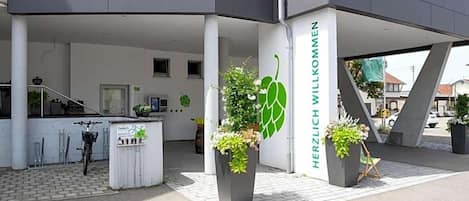 Une entrée moderne, agrémentée d’une pancarte accueillante, de plantes en pot et d’un vélo garé à l’extérieur.
