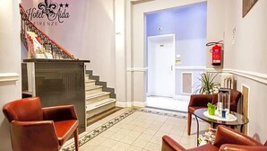 A hotel lobby with a staircase, a reception desk, leather chairs, a small table with a plant, and a fire extinguisher. - Hotel Aida (Florence)
