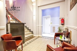 Eine Hotelhalle mit Treppe, einer Rezeption, Ledersesseln, einem kleinen Tisch mit Pflanze und einem Feuerlöscher.