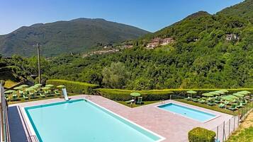 Ein Poolbereich mit zwei rechteckigen Pools, umgeben von grünen Sonnenschirmen und Liegestühlen, vor einer Kulisse aus üppigen grünen Hügeln und blauem Himmel.