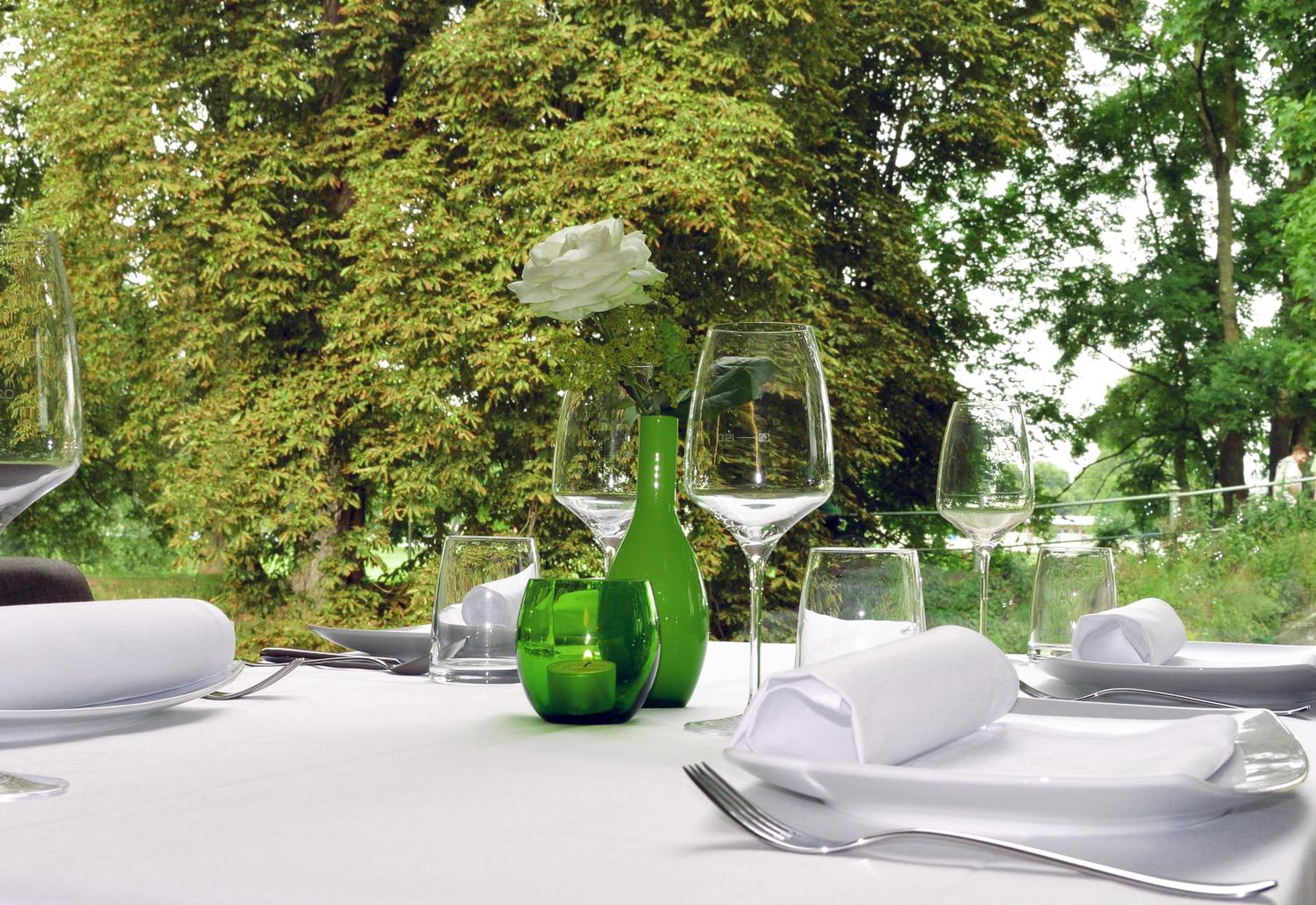 Une table dressée à l’extérieur, avec un vase vert, des serviettes blanches et des verres à vin.