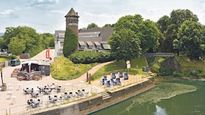 Ein Hotel mit einem Turm, Außenbestuhlung und einem Fluss.