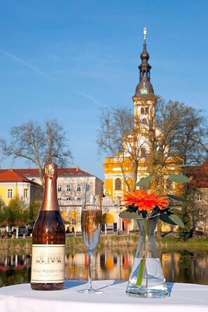 Une bouteille de vin, un verre de champagne et un vase rempli de fleurs sur une table, avec le reflet d’un bâtiment en arrière-plan.