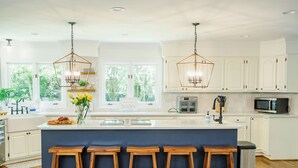 Cocina moderna con armarios blancos, una isla central con taburetes, iluminación colgante y vistas a zonas verdes.