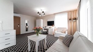 Habitación de hotel con alfombra estampada, sofá blanco, una mesita con un florero, escritorio con una silla, televisor y armario.