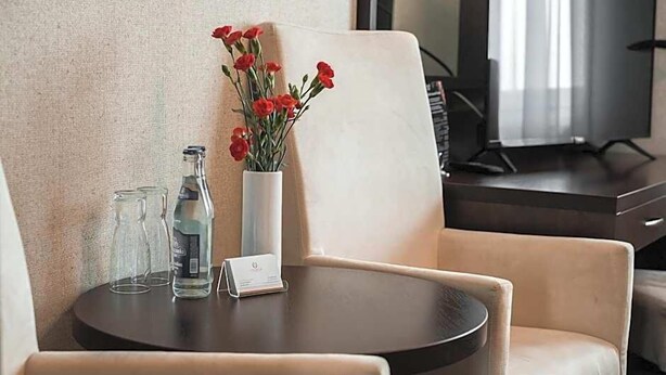 Habitación de hotel con una mesa redonda, dos sillas, un florero con flores rojas, una botella y un vaso.