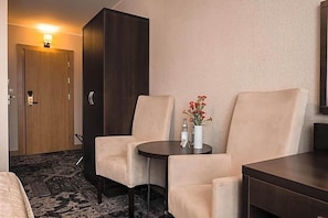 A hotel room with a bed, a dark wooden wardrobe, two beige armchairs, a small round table with a vase of flowers, and a wall-mounted TV. - Hotel Coloseum (Jaroslaw)